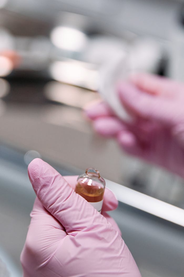 Detailed close-up of hands wearing pink gloves holding a medicinal ampoule in a laboratory setting.